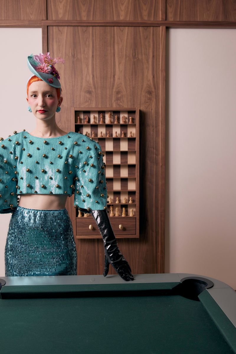 Woman in turquoise hat and sequin outfit poses at a pool table in an elegant room with wood paneling and a chess set on the wall.