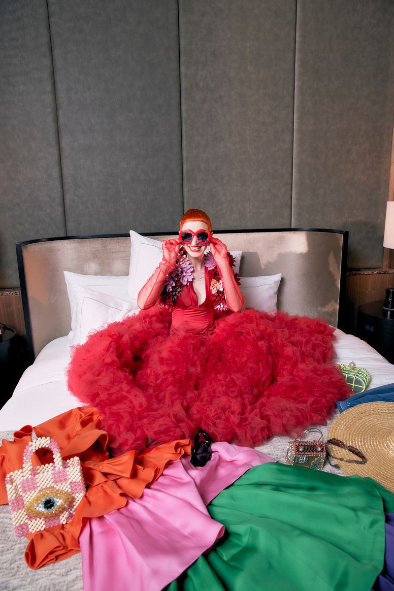 Elegant woman in a red tulle gown and heart-shaped sunglasses sits on a bed surrounded by colorful clothing and accessories.