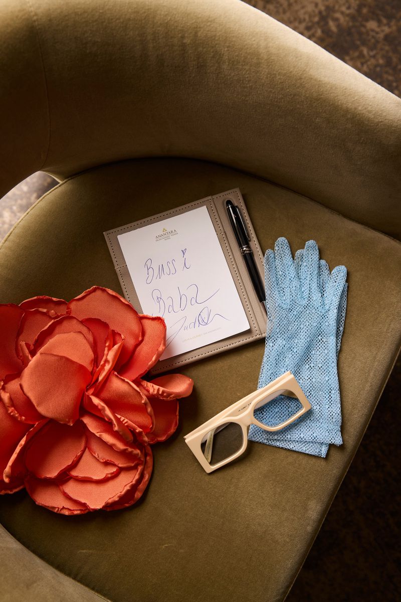 Still life on olive velvet chair: peach flower, notebook, pen, white glasses, blue lace gloves. Warm tones, close-up.