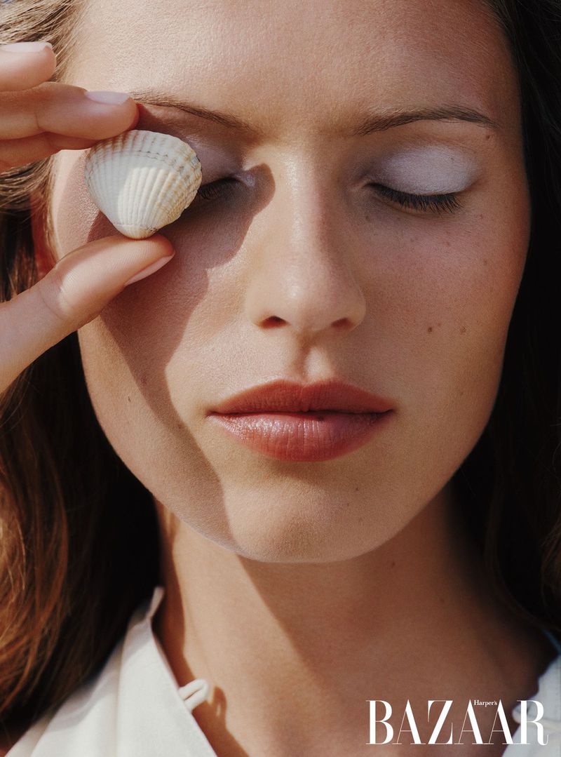 Close-up portrait of a woman with fair skin, eyes closed, holding a shell over one eye, with 'BAZAAR' logo visible at the bottom.