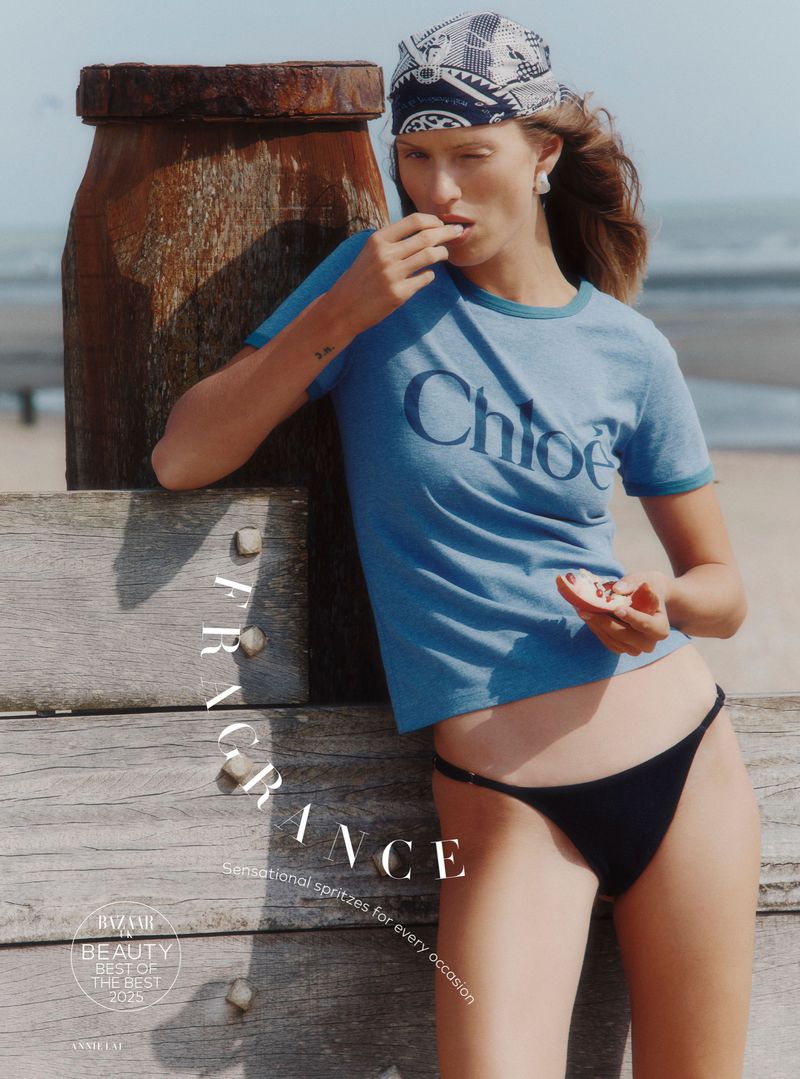 Woman in branded blue tee and black bikini eating a fruit on a beach, with wooden pier and water visible in the background on a sunny day.