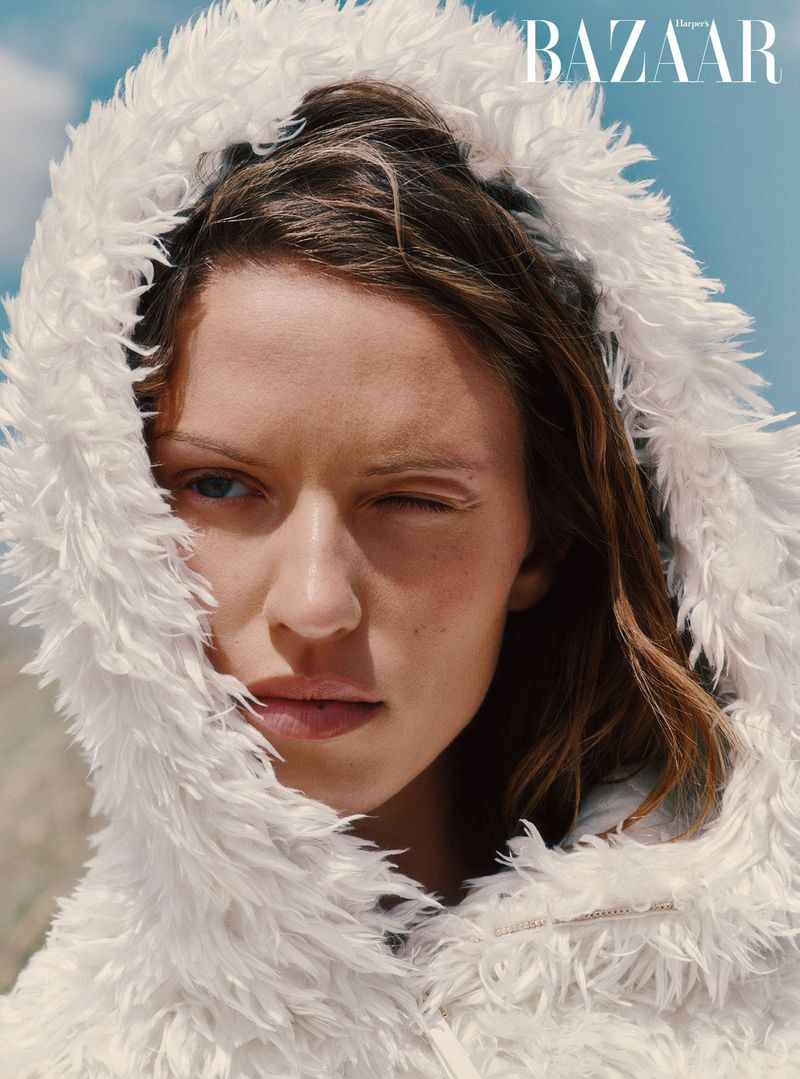 Close-up portrait of an adult female winking, wearing a white furry hooded jacket, against a blue sky. BAZAAR magazine logo visible.