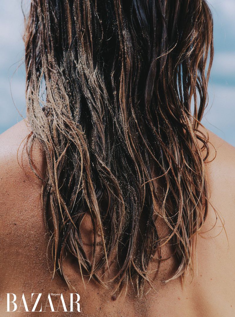 Close-up shot of long wet brown hair with sand on a person's back, set against a blurred blue background and the 'BAZAAR' logo.