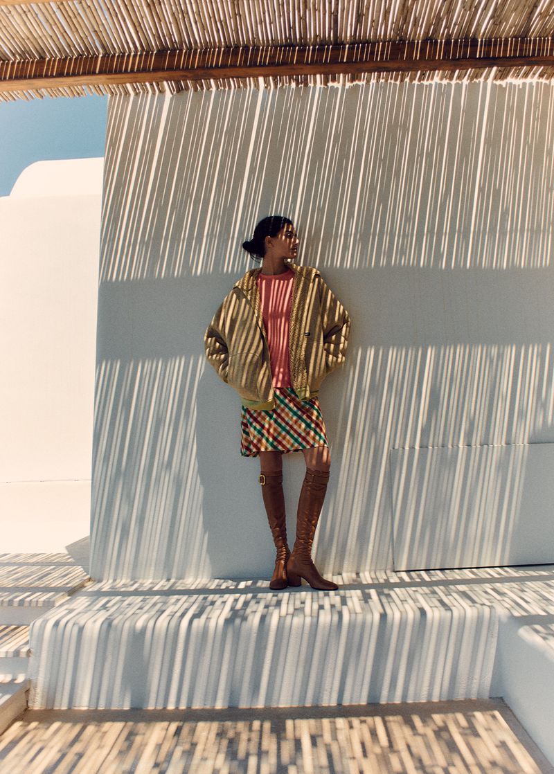 Woman in a striped top, plaid skirt, and knee-high boots poses against a white wall with strong shadows from a reed canopy above.