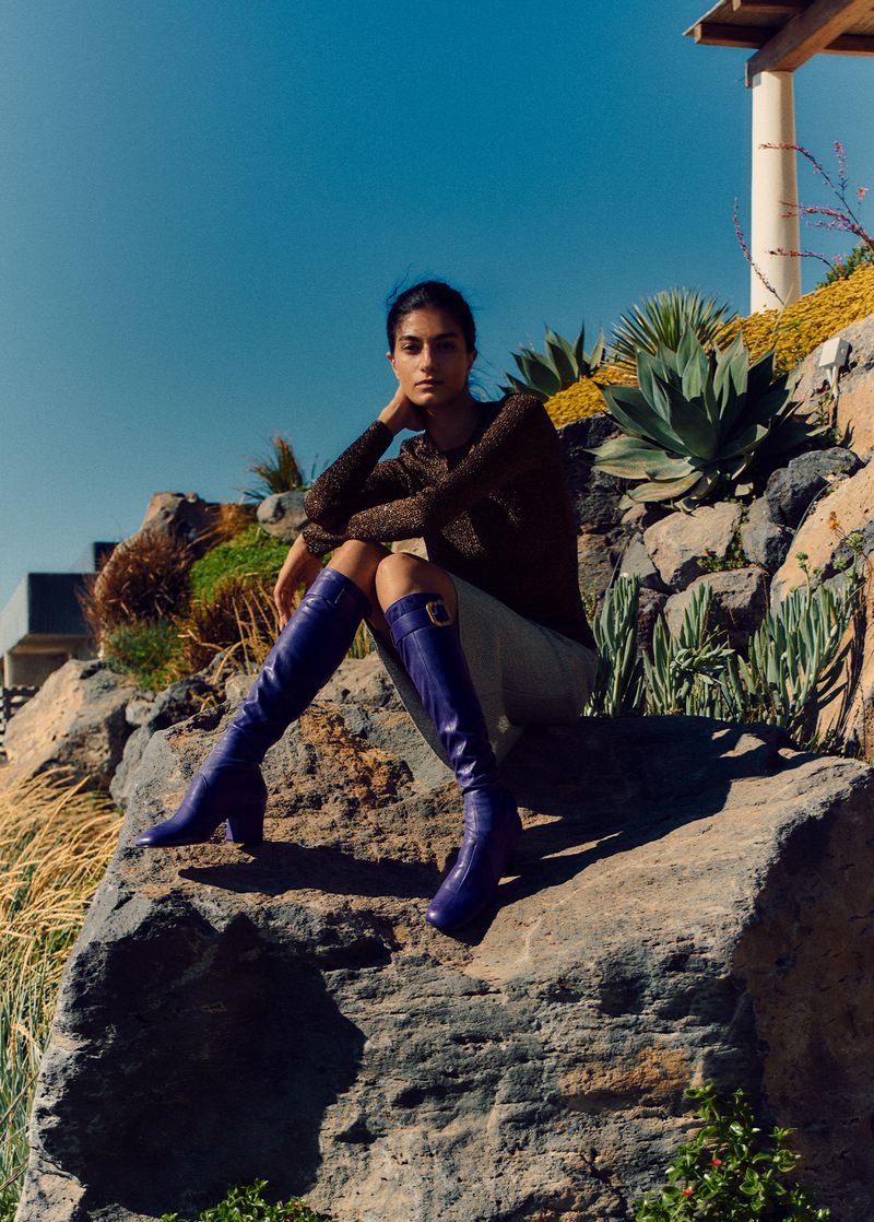 Brunette woman in a sparkling top and knee-high purple snakeskin boots sits on a rock formation in a sunny outdoor setting.