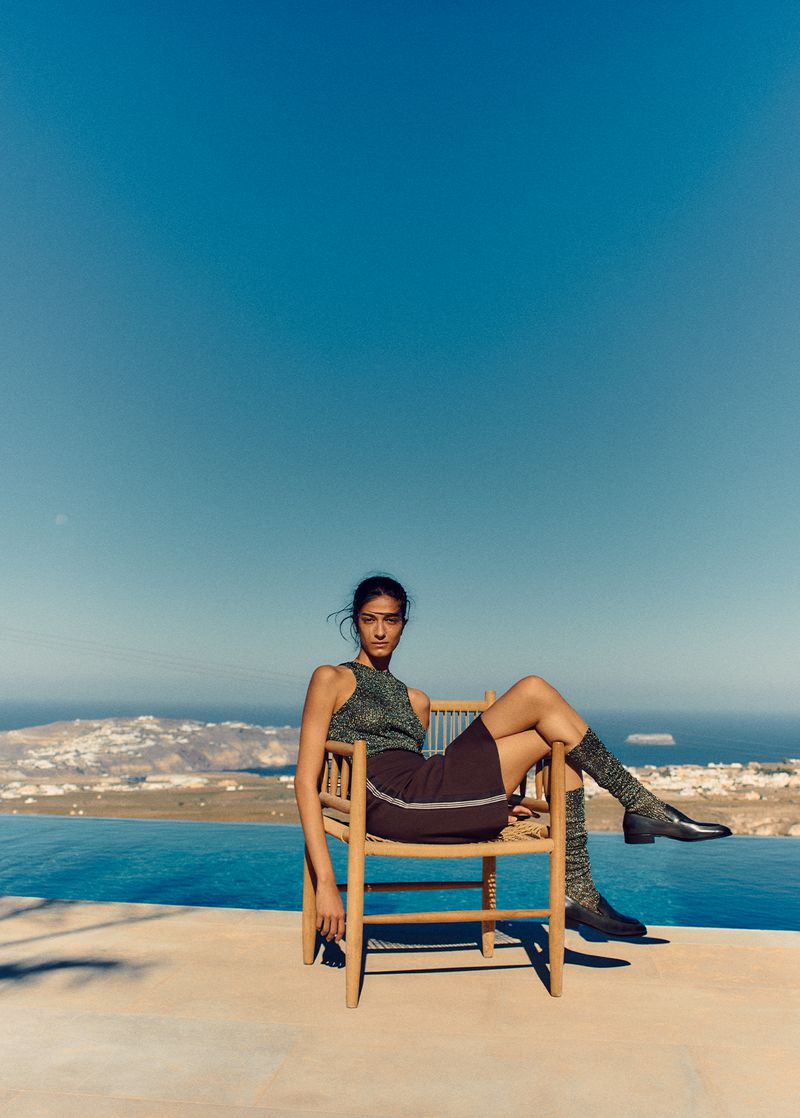 A woman in a sparkling top and skirt, sitting on a chair on a terrace with a view of the water and sky.