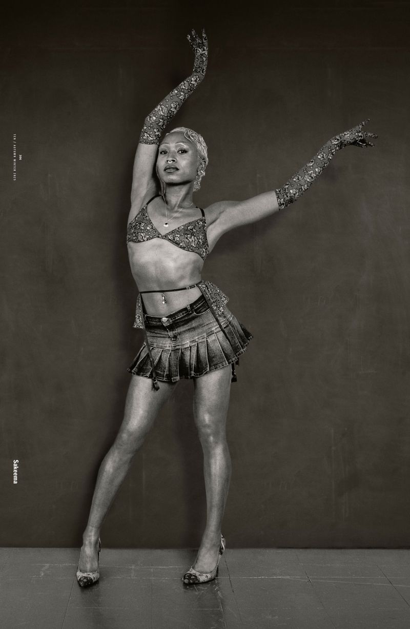Monochrome studio portrait of an androgynous adult with silver hair, wearing a bra top and denim skirt, with arms raised and wearing gloves.