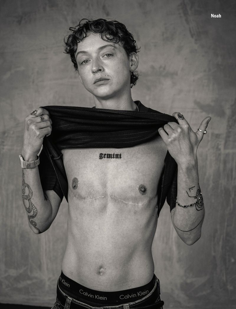 Portrait of a shirtless person with the word 'Gemini' tattooed on their chest, wearing jeans and bracelets against a gray background.