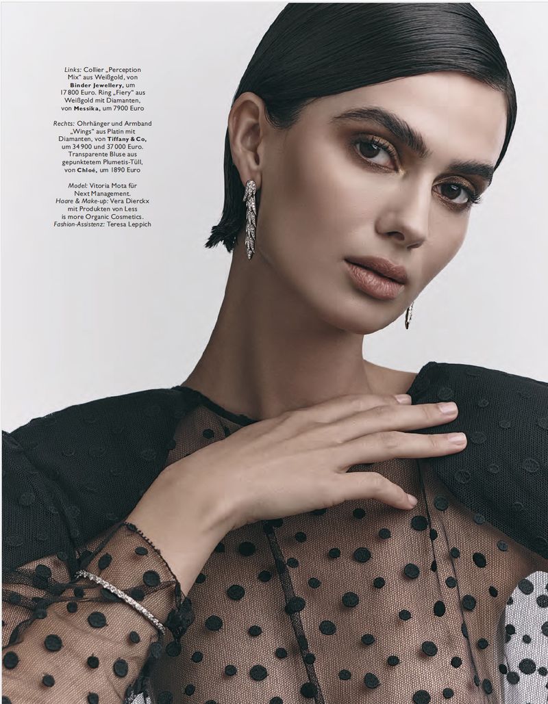 A brunette woman poses in a sheer black polka dot top with jewelry, in a studio shot with soft lighting and a neutral background.