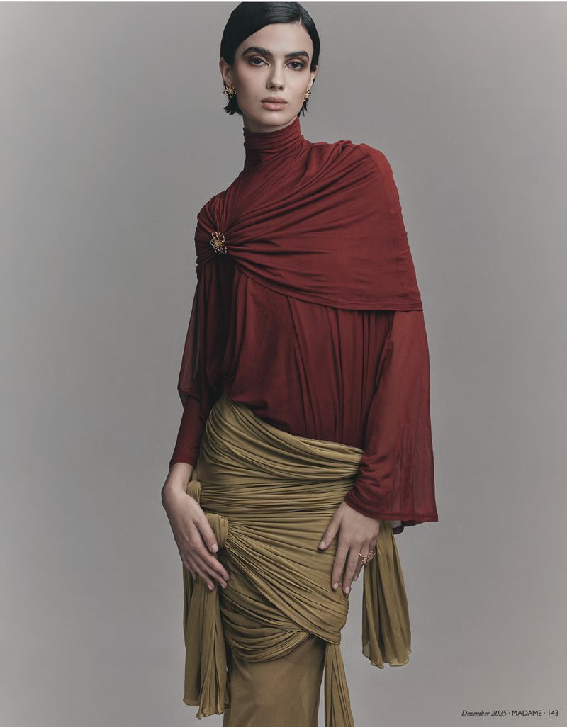 A woman poses in a red turtleneck and olive green draped skirt against a neutral backdrop in a fashion editorial shot, looking at the camera.