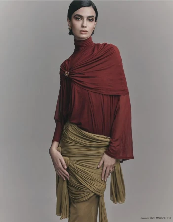 A woman poses in a red turtleneck and olive green draped skirt against a neutral backdrop in a fashion editorial shot, looking at the camera.