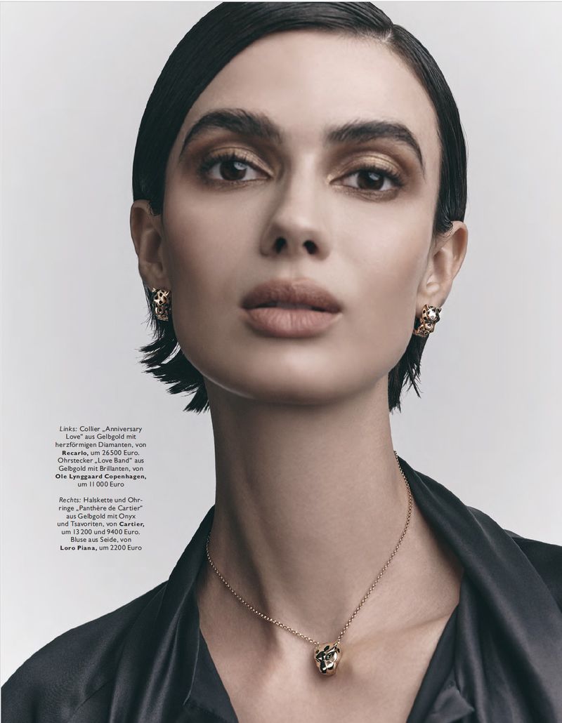 Close-up of a woman with short, dark hair wearing gold jewelry, including earrings and a necklace with a gold pendant.