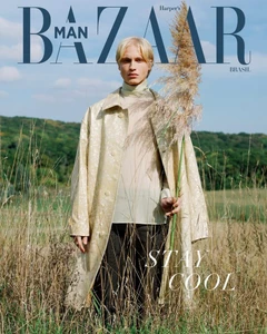 Blonde male model wearing a light beige patterned coat standing in a grassy meadow under a clear sky.