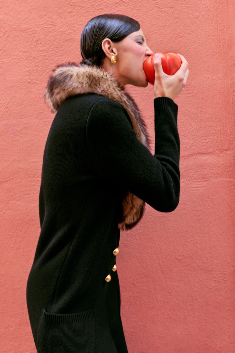 Elegant woman in a black cardigan with fur collar bites into a red tomato against a coral-colored wall, showcasing a blend of fashion and food.
