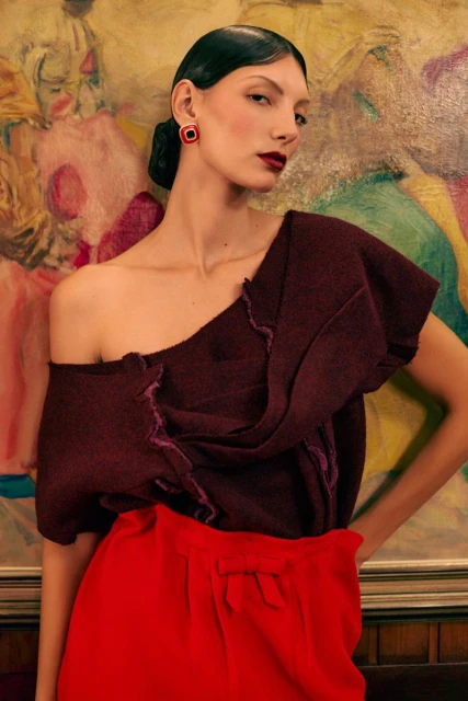 Fashion shot of a female model in a maroon, one-shoulder top and bright red pants posing against a backdrop of an abstract painting.