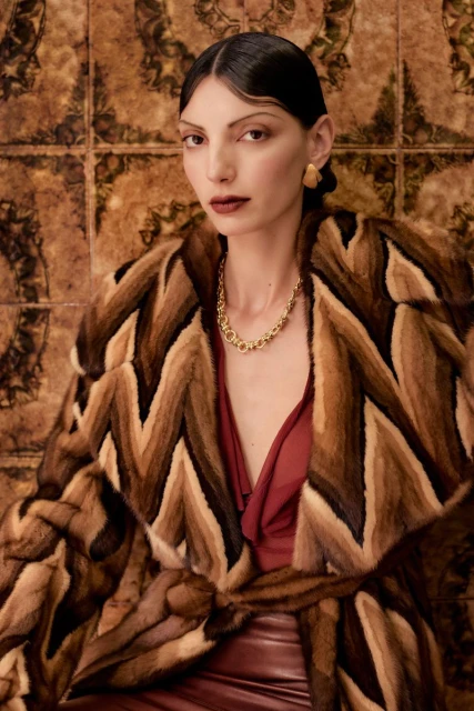 Brunette woman with neutral expression wearing statement gold necklace and patterned fur coat against a tiled background.