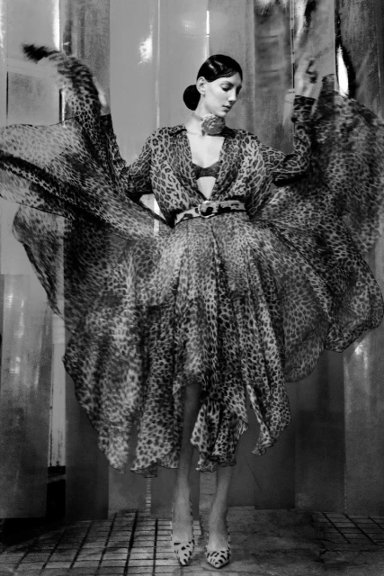 A woman poses in a dramatic leopard-print dress with matching high heels against a textured, neutral backdrop in a fashion editorial.