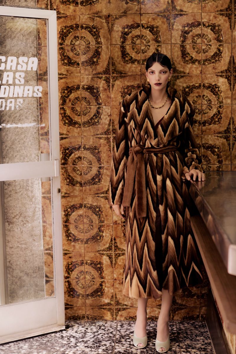 Elegant adult female in a patterned brown coat and light-green high heels poses against a detailed geometric tile wall and marble countertop.