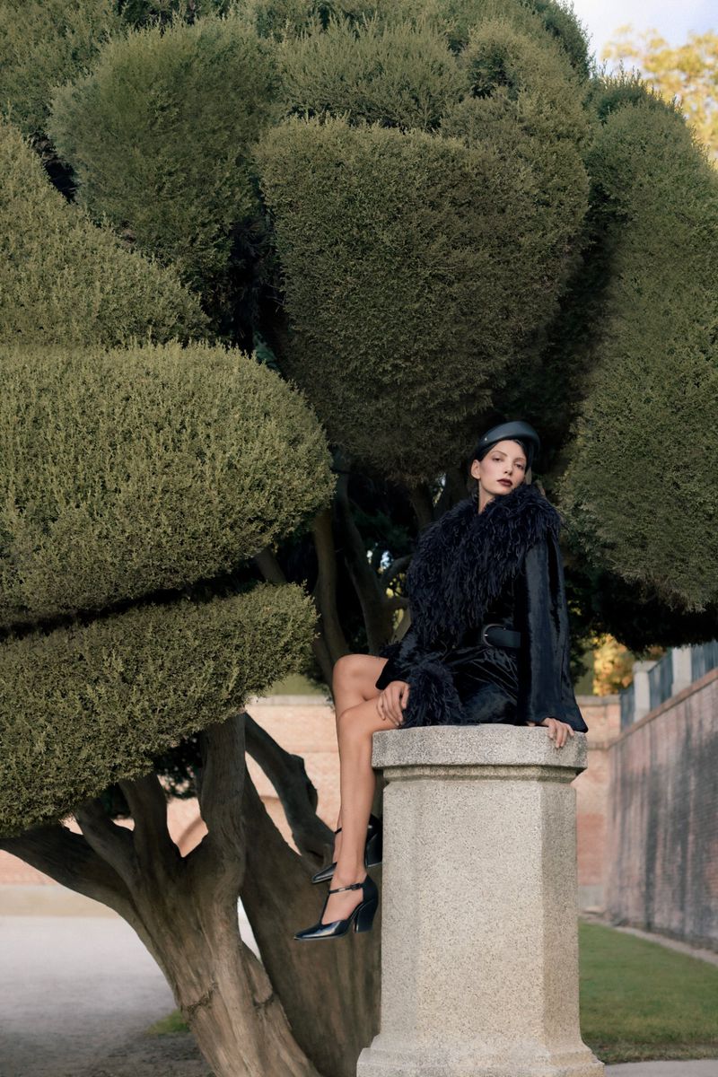 Adult female model sits on stone pedestal in stylish black dress, heels, and hat, accessorized with fur scarf, amid leafy trees in park.