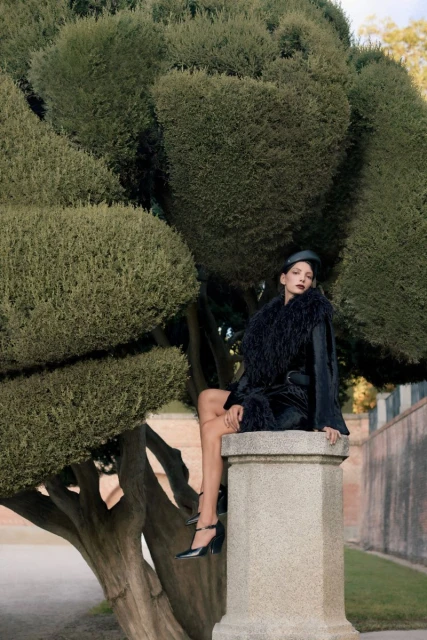 Adult female model sits on stone pedestal in stylish black dress, heels, and hat, accessorized with fur scarf, amid leafy trees in park.