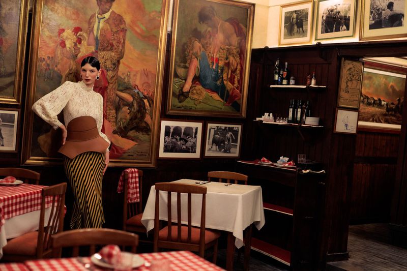 An adult woman wearing a patterned skirt and white lace top poses in a restaurant with paintings and red and white tablecloths.