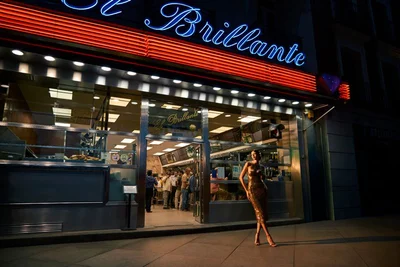 Model wearing a gold sequin dress standing outside a neon-lit restaurant at night for Harper's Bazaar editorial.