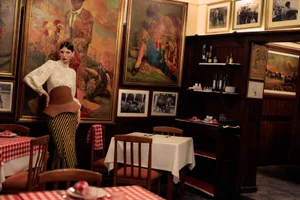 Model Cynthia Arrebola poses in a cream lace blouse and striped skirt inside a traditional dining room with framed art.