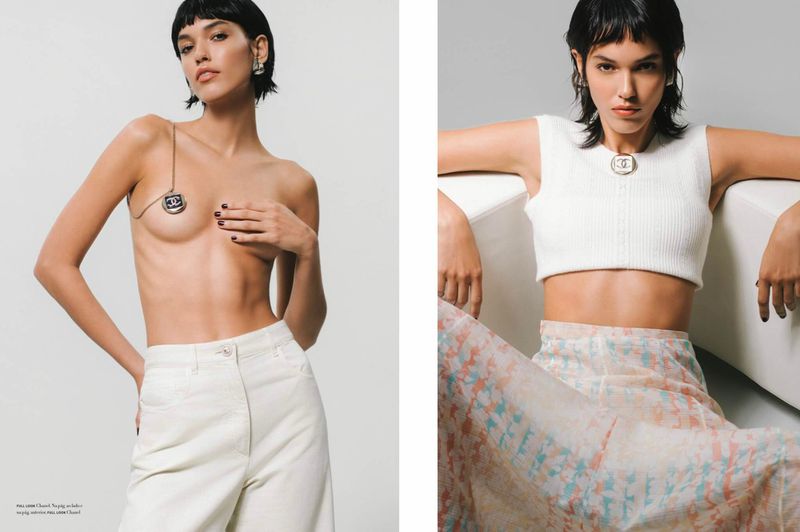 Fashion editorial featuring a model in white pants and a crop top, accessorized with a designer logo chain and earrings, set against a light grey backdrop.