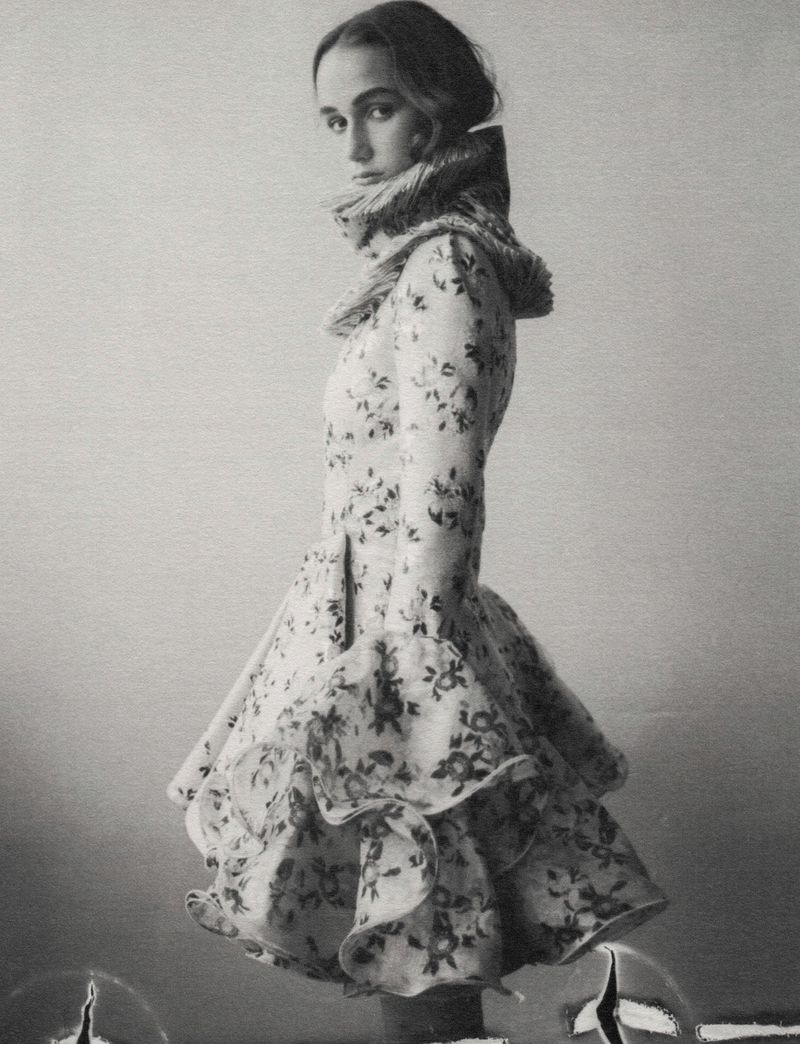 Monochrome portrait of a slender young woman in a vintage-style floral coat with a furry collar, posing in a photo studio setting.