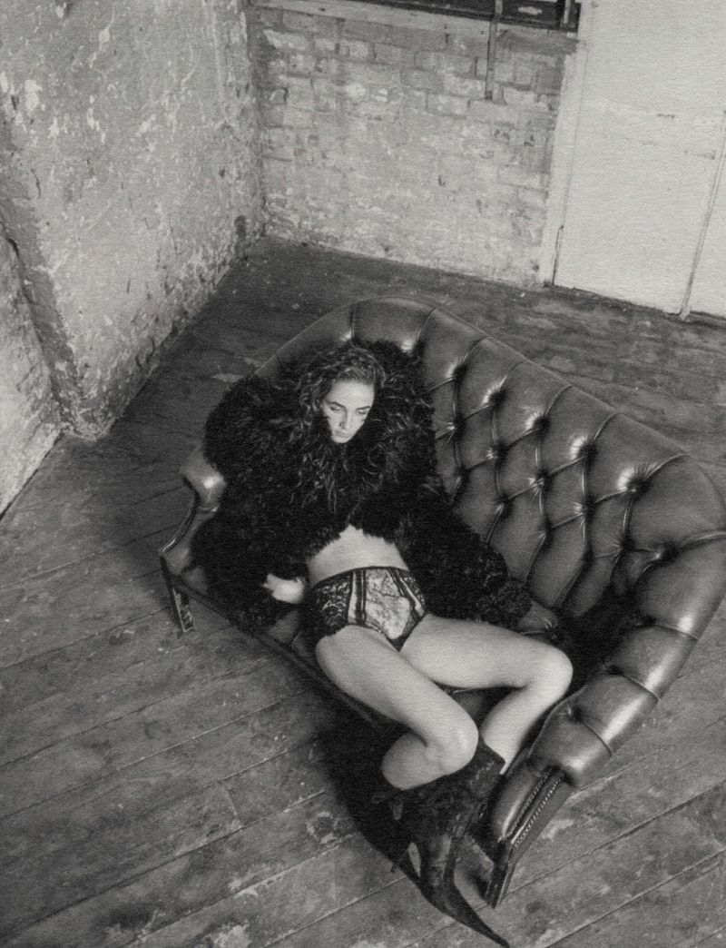 Black and white portrait of a woman lying on a leather sofa wearing a fur coat and underwear in a sparse room with distressed walls.