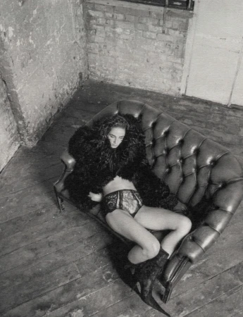 Black and white portrait of a woman lying on a leather sofa wearing a fur coat and underwear in a sparse room with distressed walls.