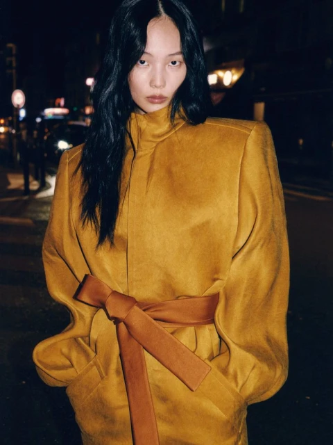 Stylish woman in mustard coat with brown sash poses at night with blurred street lights and pedestrians in background. Fashion, beauty.