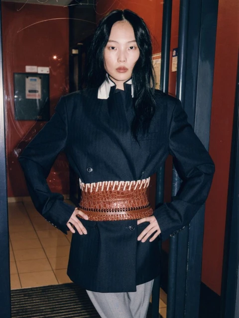 Adult female fashion model posing in a dark blue suit jacket and brown waist cincher belt indoors against a dark red wall.