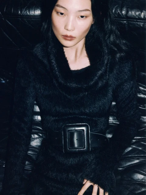 A long shot of a single adult female model with dark hair wearing a dark faux fur coat with a large belt lying on a black leather couch.