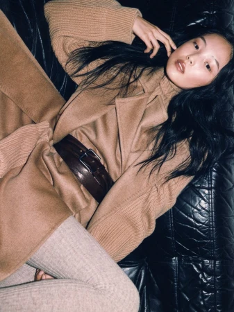 Elegant Asian woman poses on black leather couch in a tan sweater, belted coat, and leggings in a high-angle fashion portrait.