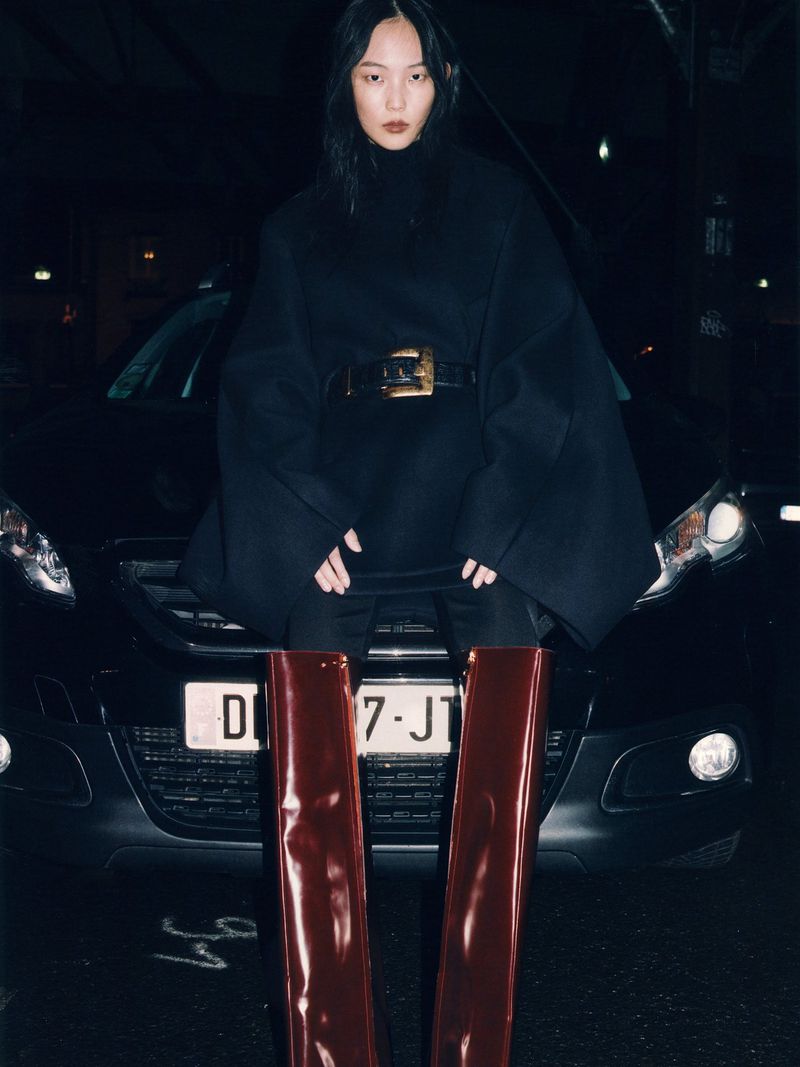 Asian female fashion model poses in a black top with large belt, patent leather thigh-high boots, and leans against a black car at night.