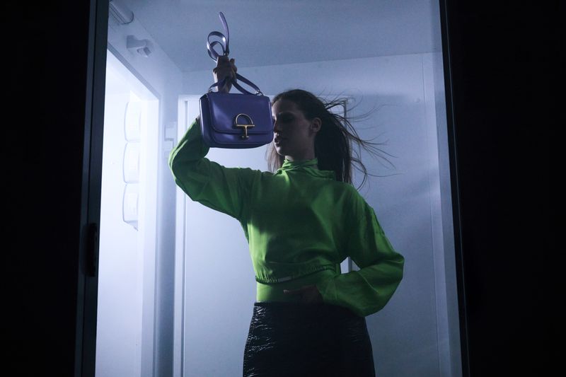 Indoor shot of a woman in green top holding up a purple satchel purse in front of a white refrigerator for a fashion editorial.