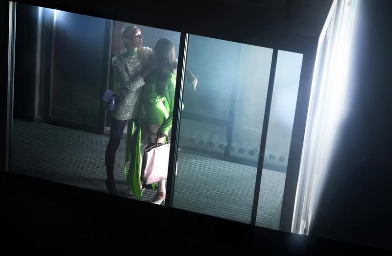 Two fashion models in glittering dresses pose indoors near a glass window with diffused lighting in a glamourous fashion editorial.