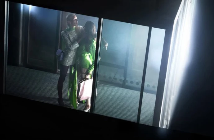 Two fashion models in glittering dresses pose indoors near a glass window with diffused lighting in a glamourous fashion editorial.