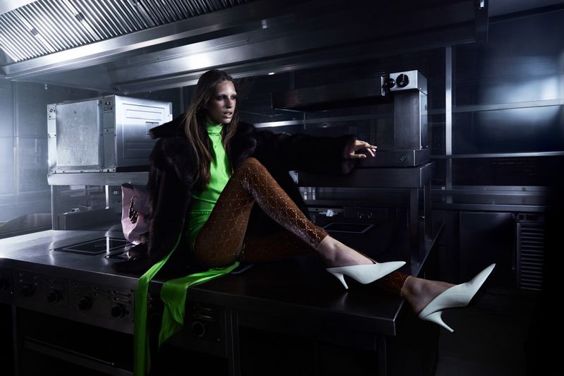 A stylish adult woman with long hair, wearing a vibrant green turtleneck, animal-print pants and white heels, poses in a commercial kitchen setting.
