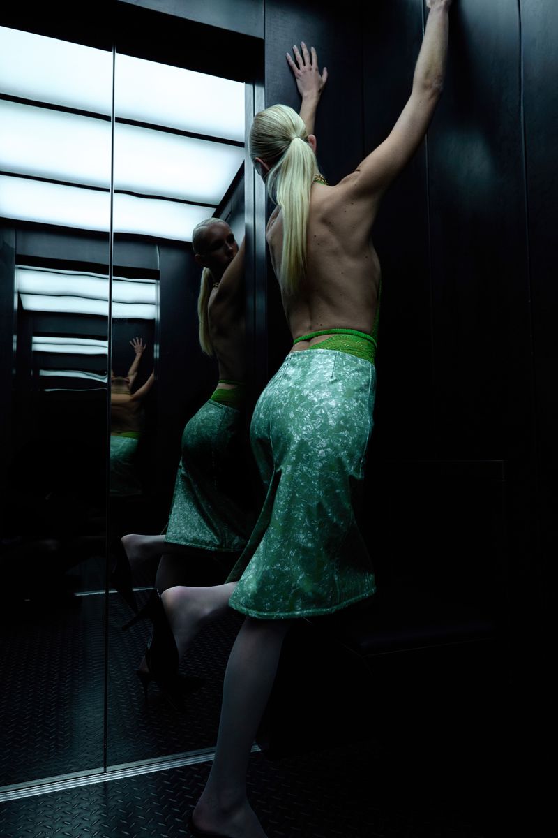 An adult female strikes a pose in a mirrored elevator. She is seen from the rear in a green skirt and is illuminated by strong overhead lighting.