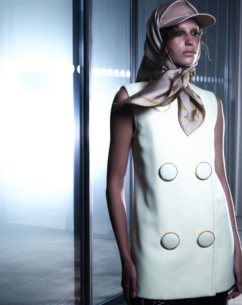 Fashionable young woman posing indoors wearing a sleeveless dress with oversized buttons, a scarf, and a baseball cap.