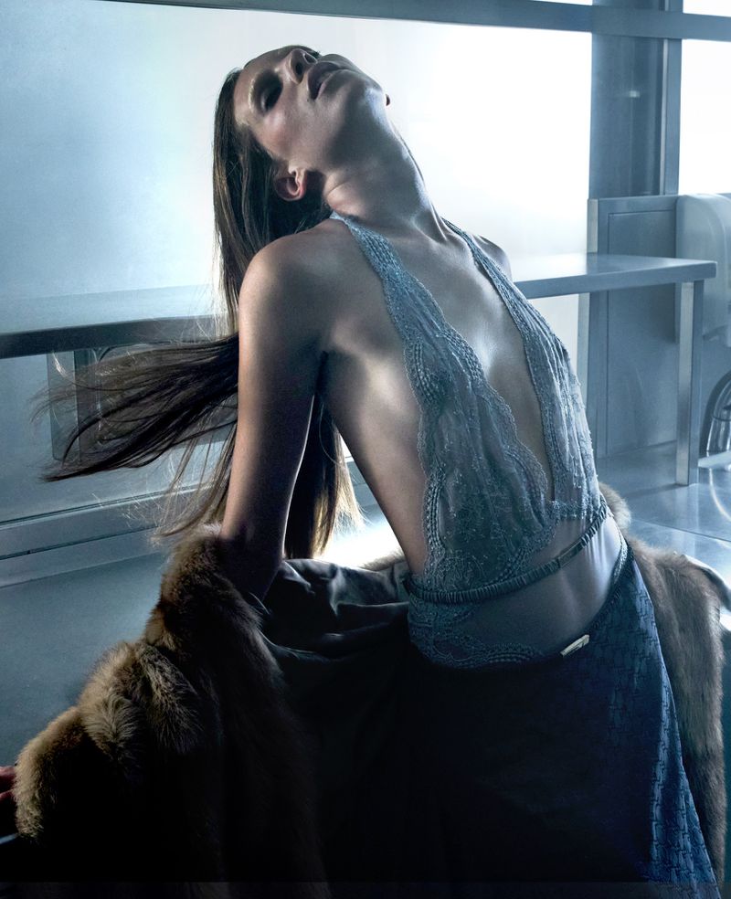 A tanned, slender adult woman tilts her head back, showcasing a light silver top with a fur stole in a studio setting with metallic accents.