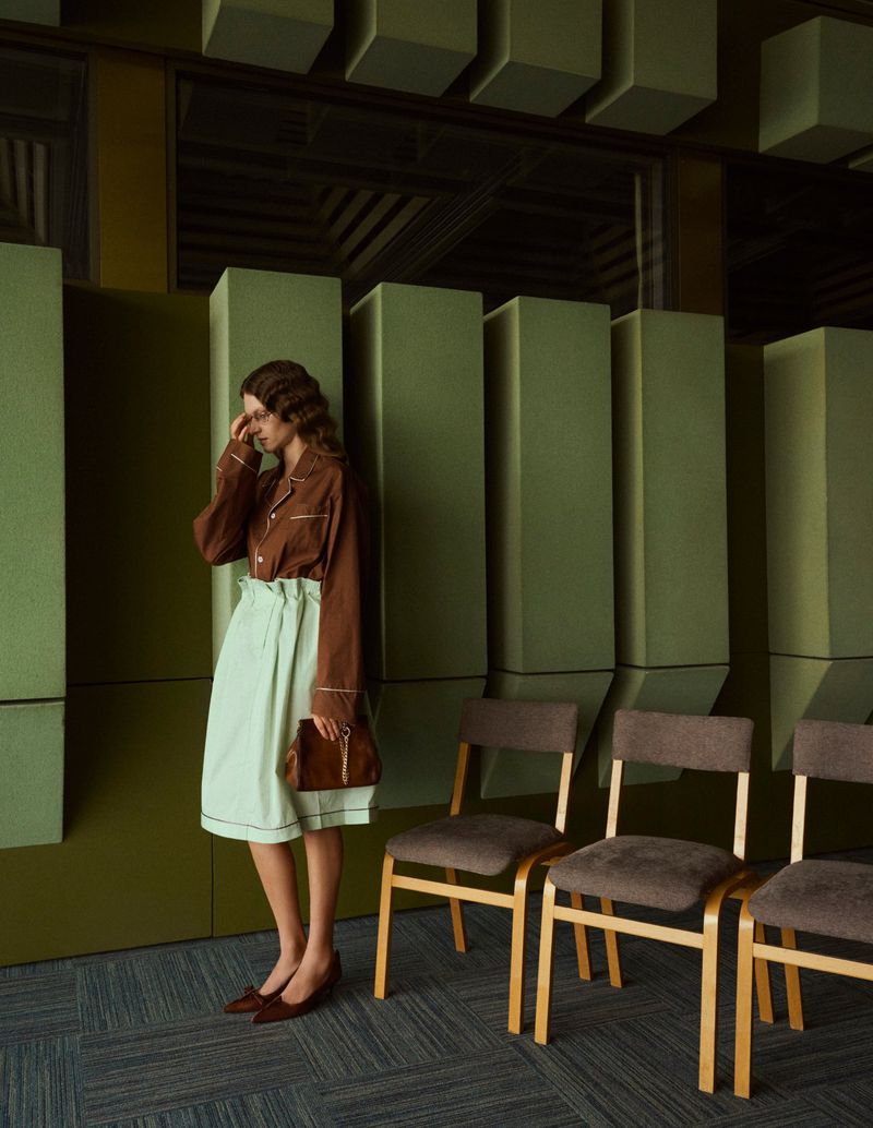 Fashion shot of a woman in a brown and mint green outfit, standing next to a row of chairs in a room with green walls and carpeted floor.