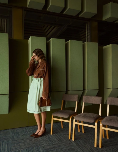 Fashion shot of a woman in a brown and mint green outfit, standing next to a row of chairs in a room with green walls and carpeted floor.