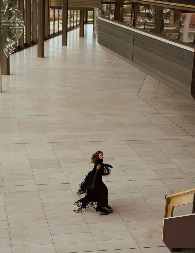 A woman in a fringed black dress stands on a marble floor inside an open-plan building featuring tall columns and large windows.