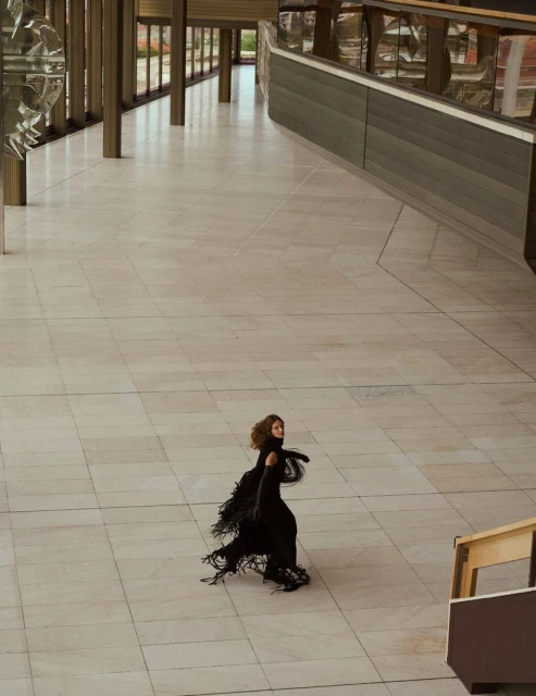 A woman in a fringed black dress stands on a marble floor inside an open-plan building featuring tall columns and large windows.