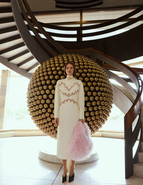 Fashion shot of a woman in an ivory dress with a pink bag, standing in front of a decorative gold sphere and spiral staircase indoors.