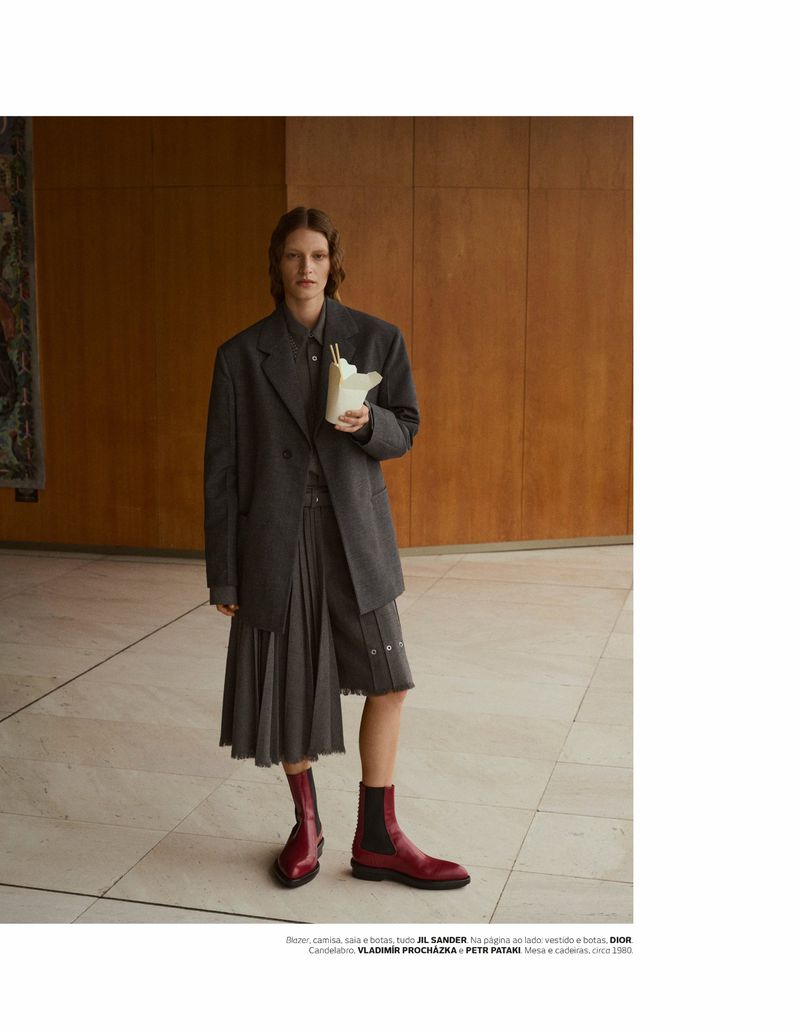 Fashion editorial shot of a non-binary person in an oversized blazer and pleated skirt, holding a white takeout container indoors.