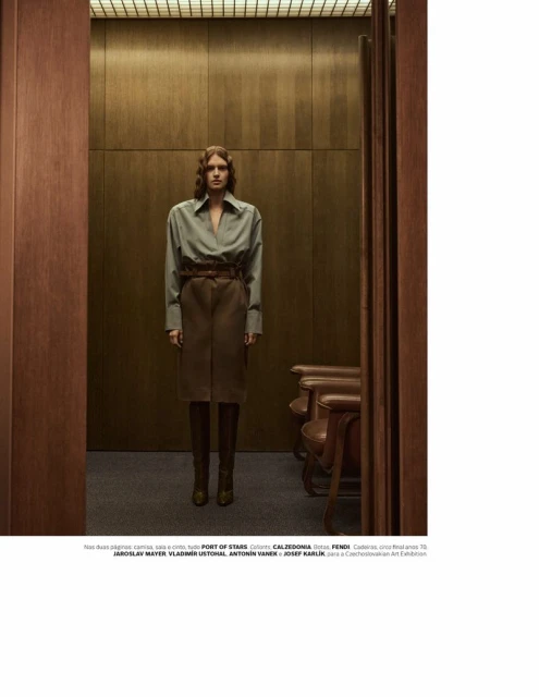 A woman in a green button-down shirt and brown skirt is standing in a doorway of a wood-paneled room with brown leather chairs.